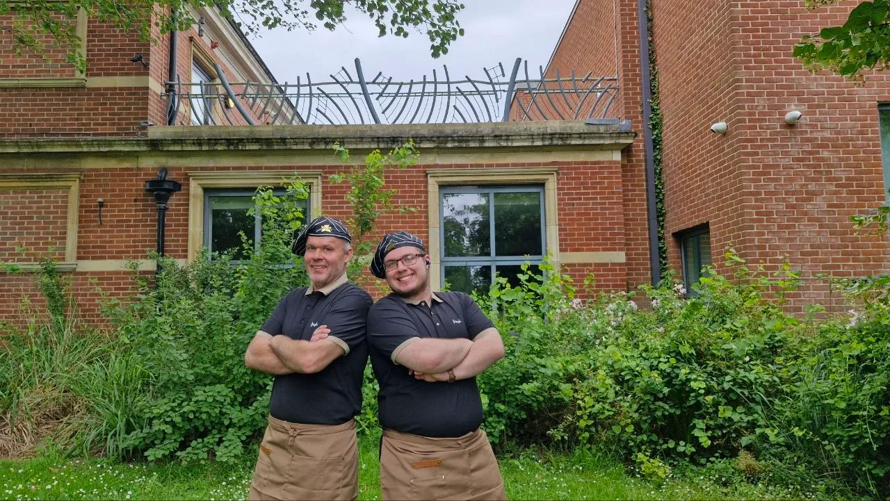 Volodymyr Kudin and his son Yehor Kudin in their workshop uniform, arms folded, outside in Hampshire
