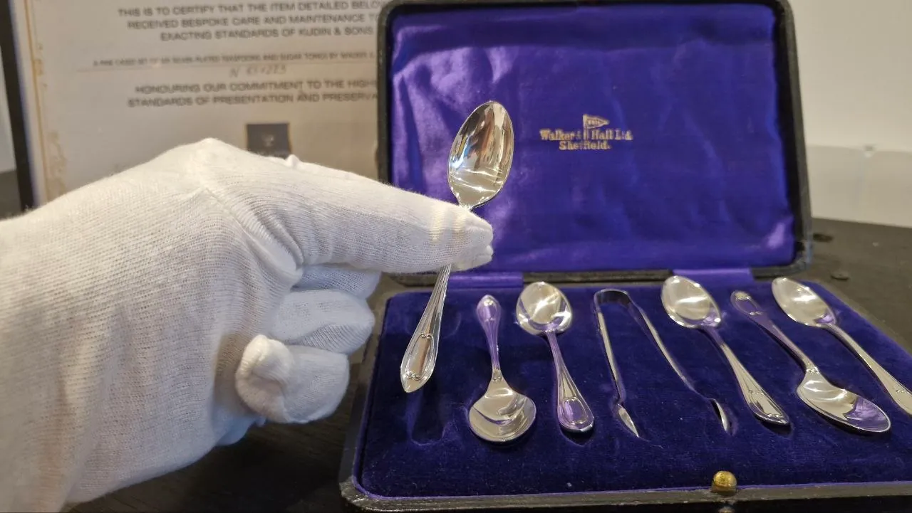 A restored Walker & Hall teaspoon held in a white cotton glove beside its case and Kudin & Sons care certificate